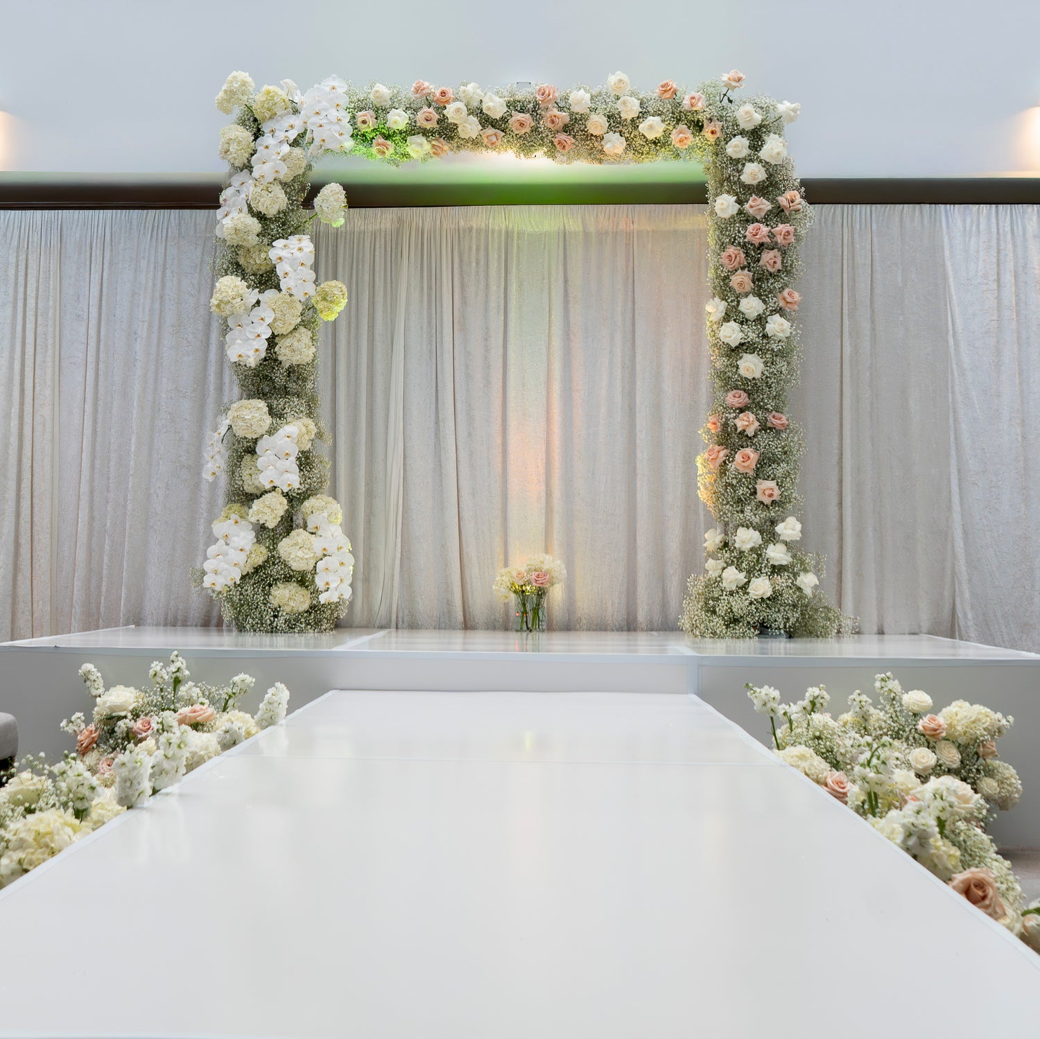 A white wedding-style event backdrop with a floral arch made of white and blush pink flowers, draped curtains behind, and a white aisle runner with matching floral arrangements along the sides.