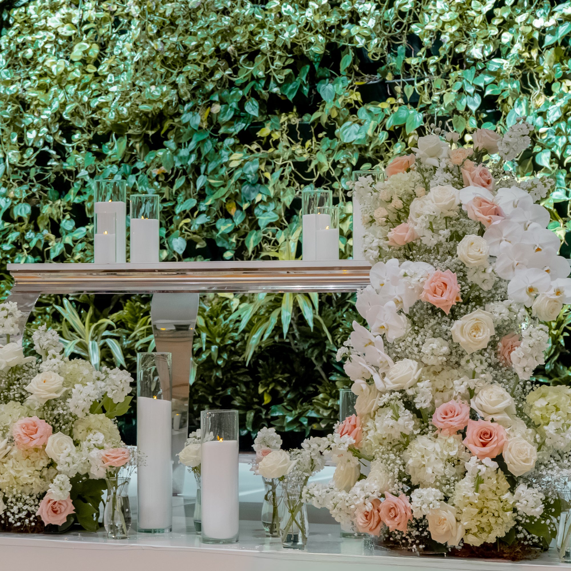 A decorative floral arrangement display with large bouquets and candles in clear glass holders, set against a lush green leafy backdrop.