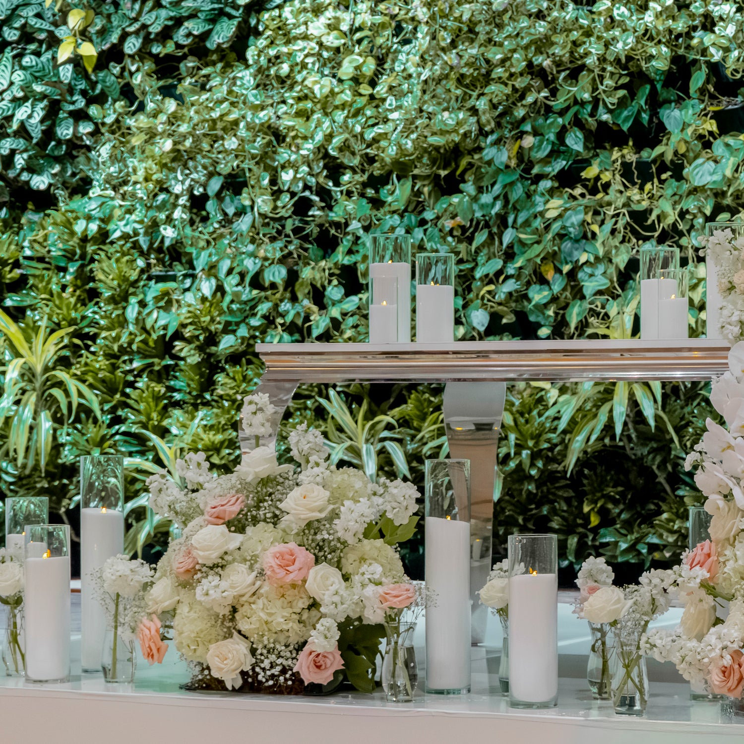 A styled floral and candle display with white and blush pink flower arrangements, clear glass candle holders, and white pillar candles set against a lush green leafy backdrop.