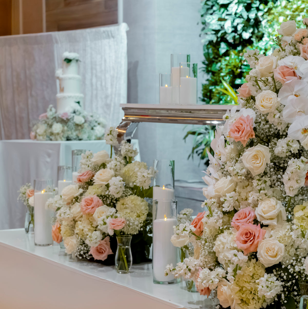 A wedding reception table decorated with large floral arrangements in white, blush pink, and cream roses, accented with white pillar candles in clear glass holders. In the background, a multi-tiered white wedding cake is displayed on a draped white tablecloth.