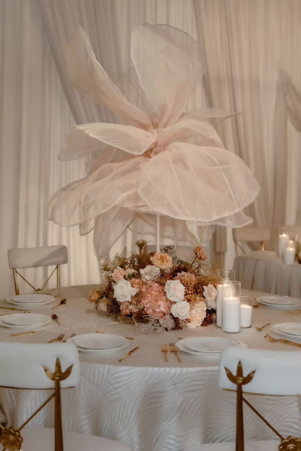 A formal event table decorated with a large pale blush tulle flower centerpiece, a floral arrangement in blush, cream, and tan tones, white plates, gold cutlery, and white draped chairs against a cream draped backdrop.