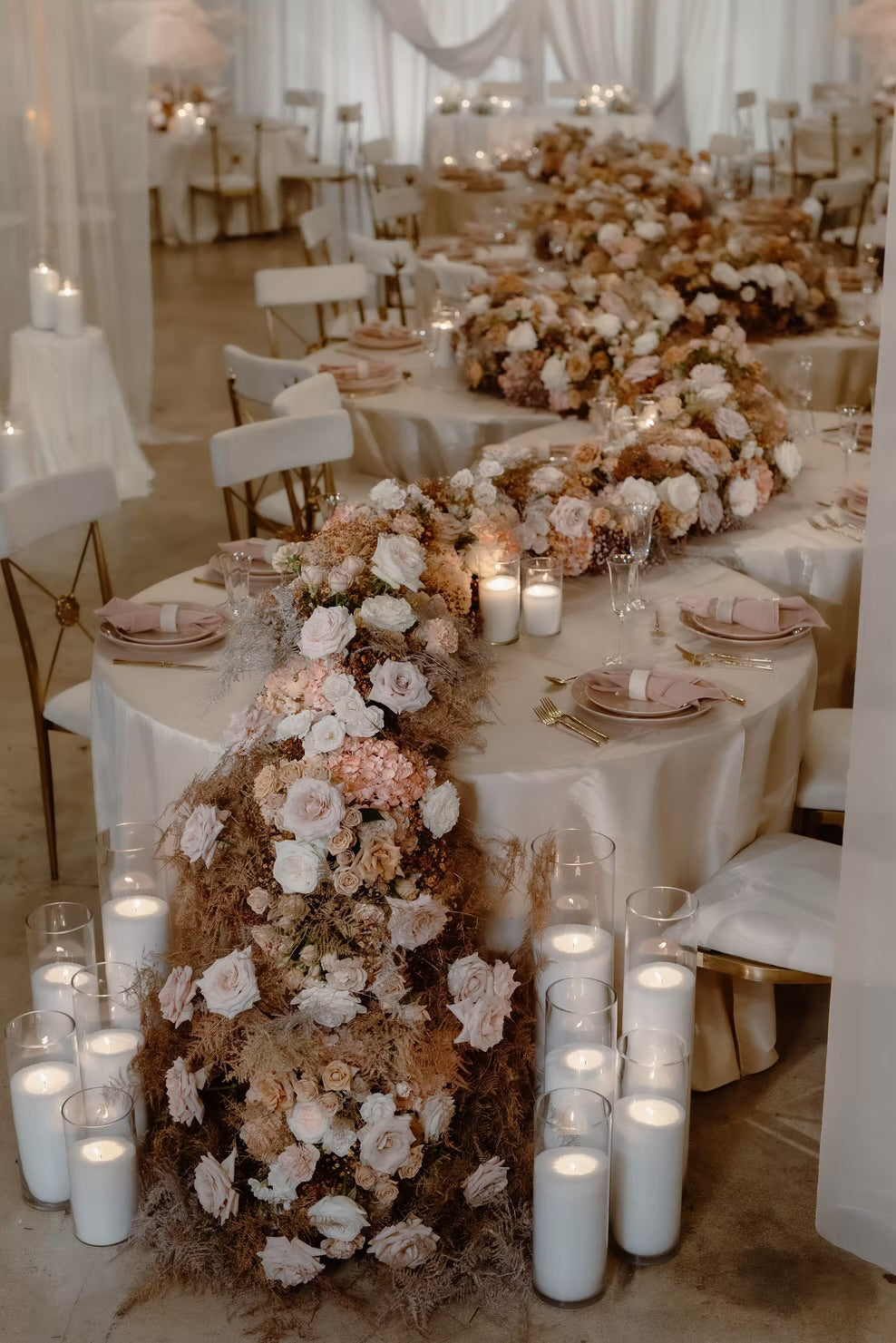 A wedding reception table decorated with a long floral centerpiece in blush pink, cream, and tan tones, surrounded by lit pillar candles in clear glass holders on a white tablecloth.