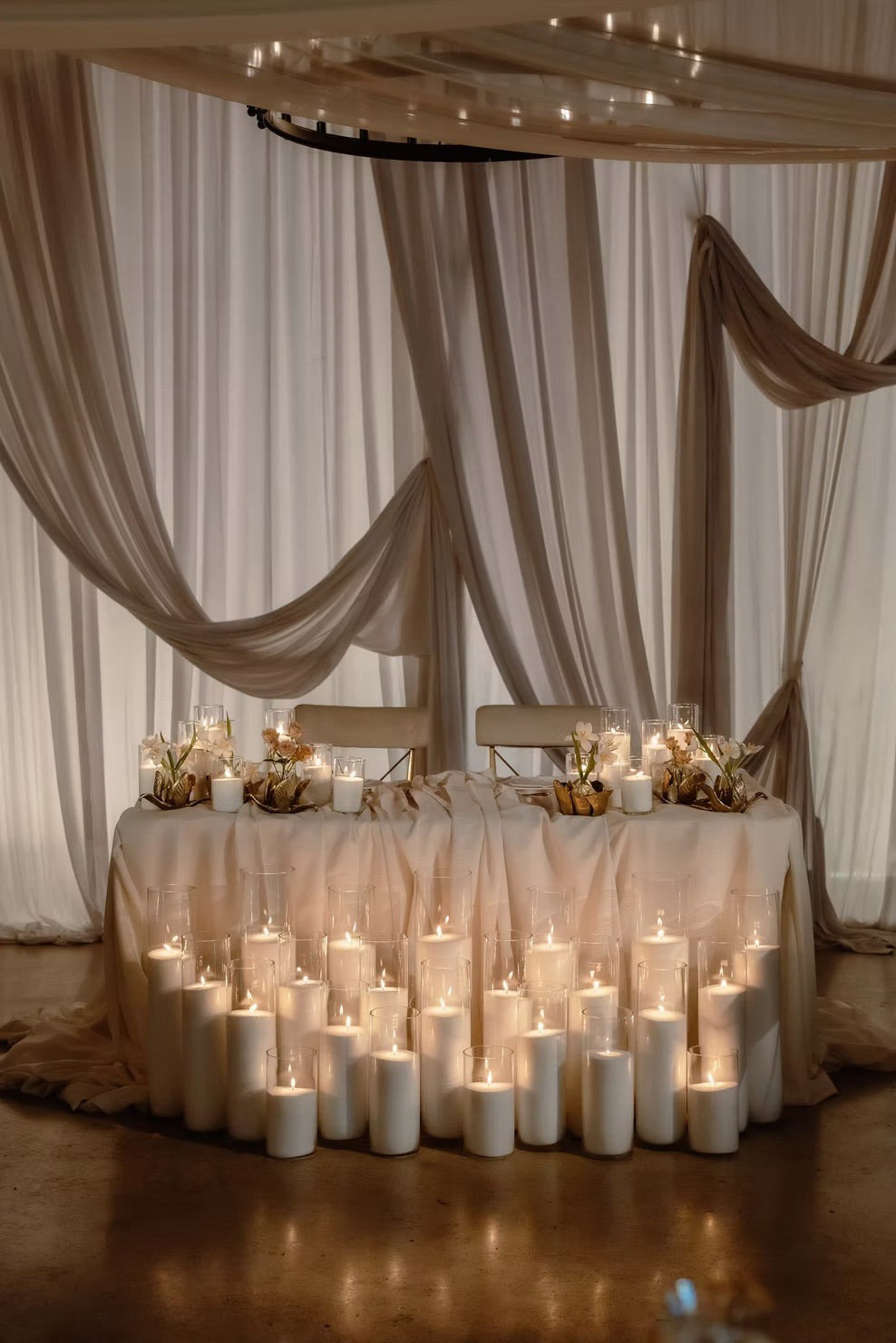 A styled wedding reception table setup with draped champagne-colored fabric, numerous lit pillar candles, and floral centerpieces under a chandelier.