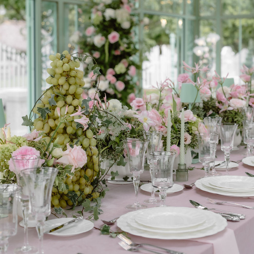 A styled event table in a bright conservatory with pale pink linens, white plates, clear glassware, and floral centerpieces including pink roses, green grapes, and leafy greenery.