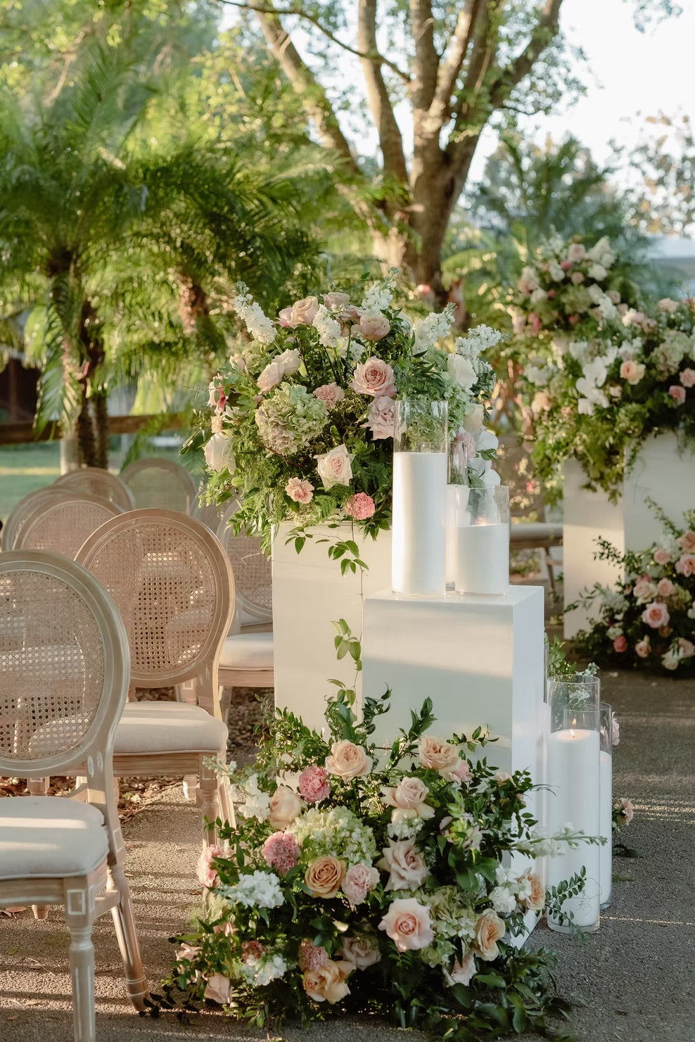 Outdoor wedding-style floral arrangements in soft pink, white, and green on white pedestals, with woven chairs and trees in the background.