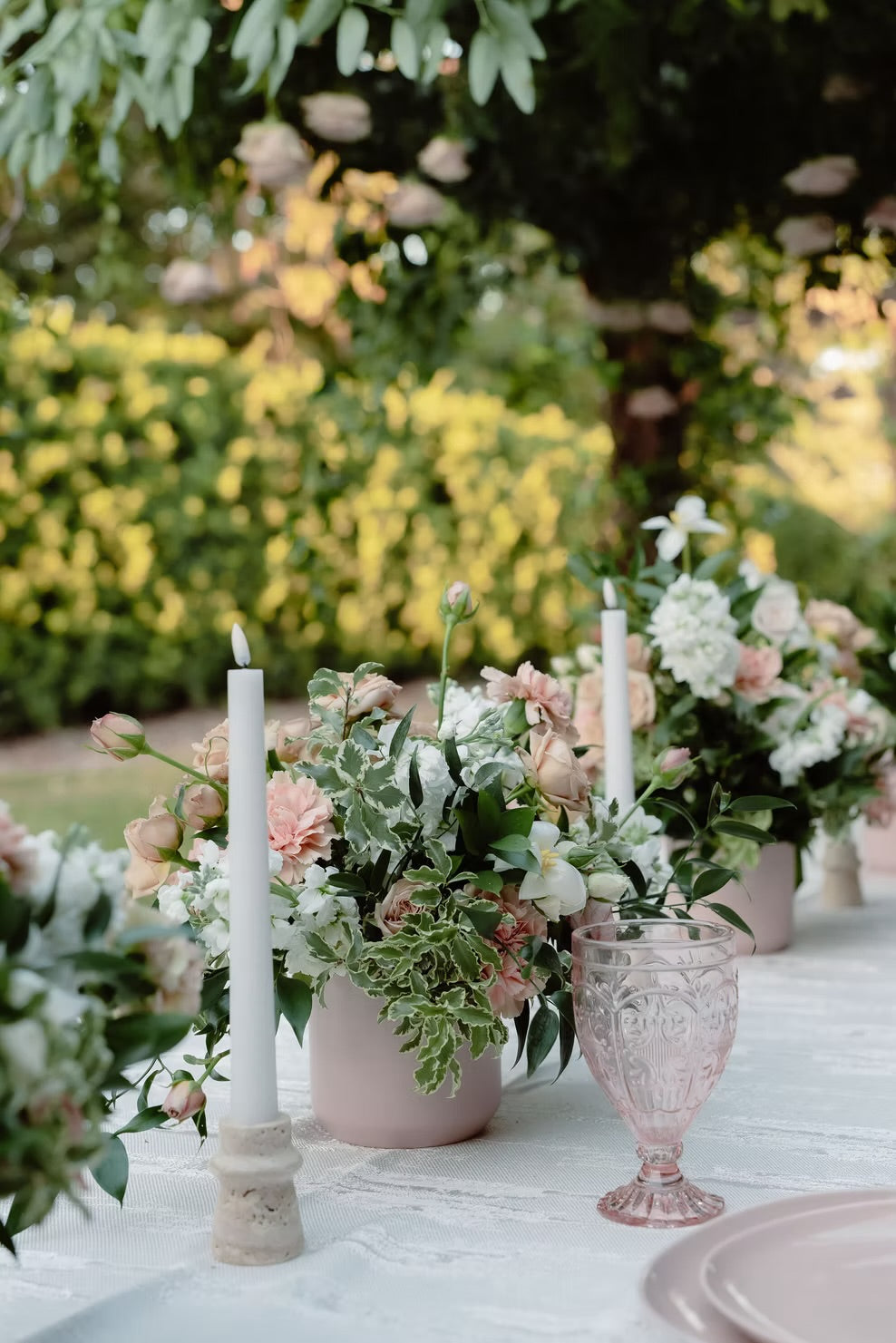 Outdoor garden table setting with pink flower arrangements in pale pink vases, white taper candles, and a pink textured glass, with a blurred green garden background.