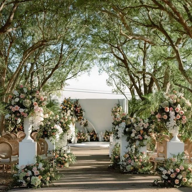 An outdoor wedding ceremony setup with a white tent backdrop, a central aisle, floral arrangements in pink, blush, and white, and rows of wooden chairs under tall trees.
