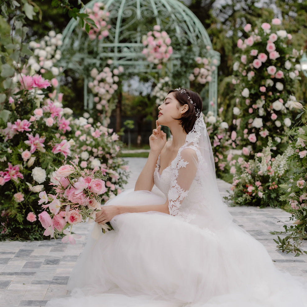 A bride in a white lace wedding gown and veil sits on a stone path in a garden, surrounded by pink and white flowers and a green arched gazebo in the background.