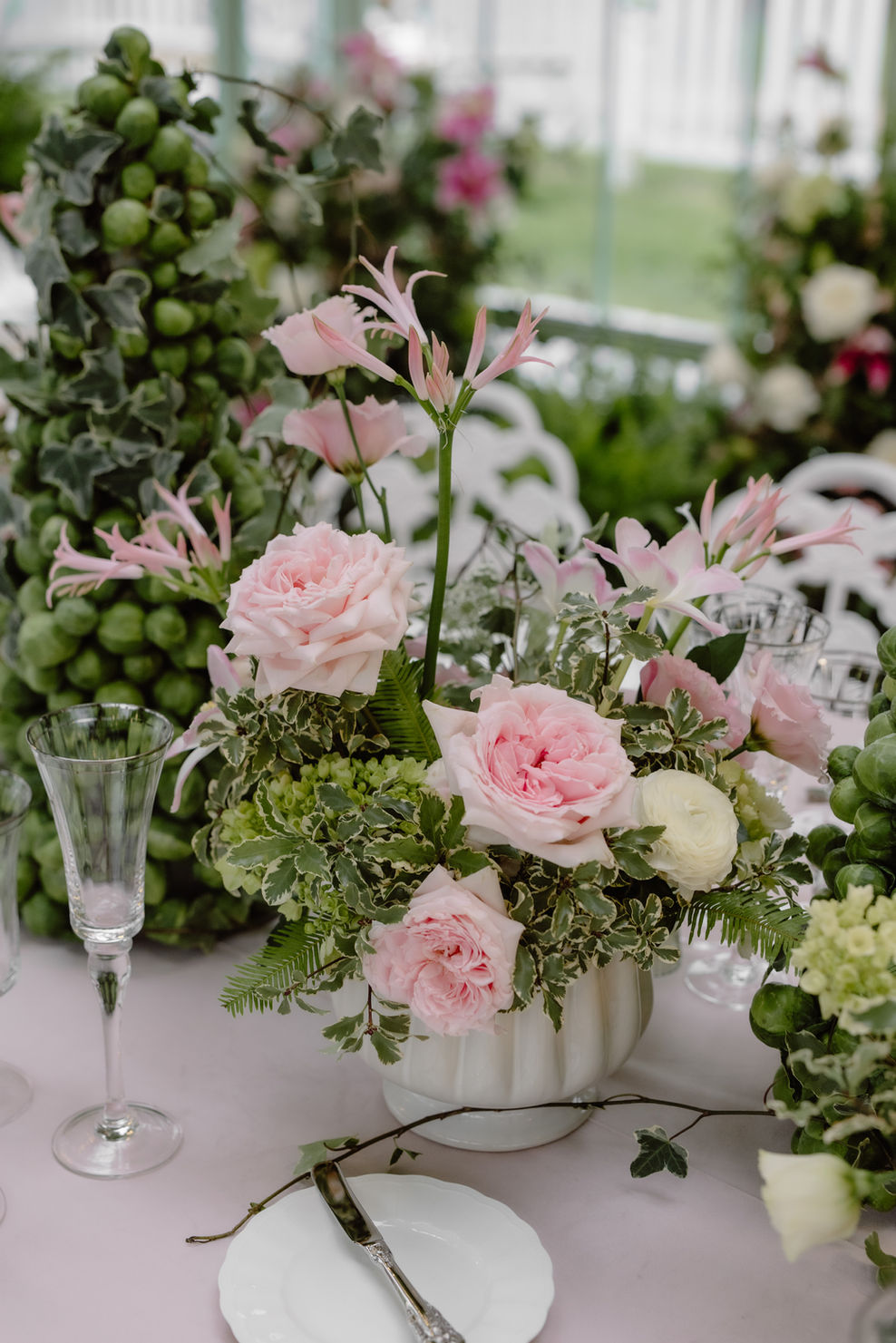 A floral centerpiece arrangement with pink roses, pale pink lilies, green foliage, and white flowers in a white ribbed vase on a table, with champagne glasses and a blurred outdoor background.