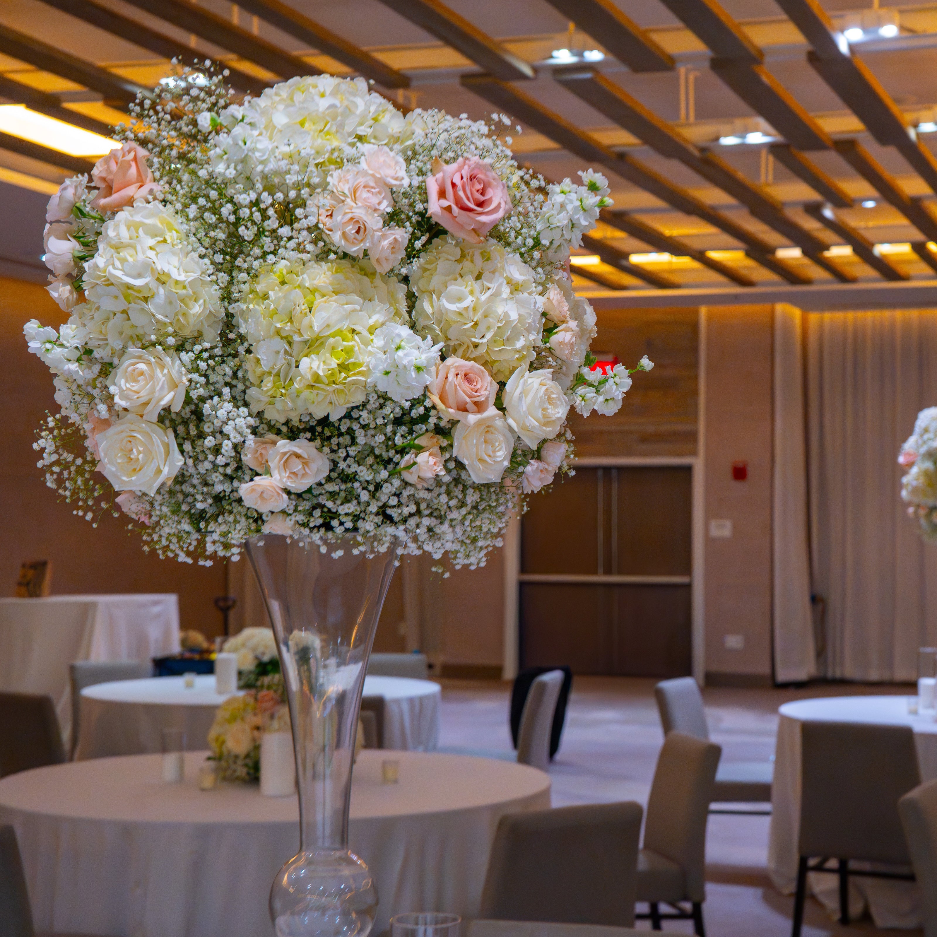 A large floral centerpiece arrangement in a tall clear glass vase, featuring white and blush pink roses with baby's breath, set in a banquet hall with round tables and chairs.