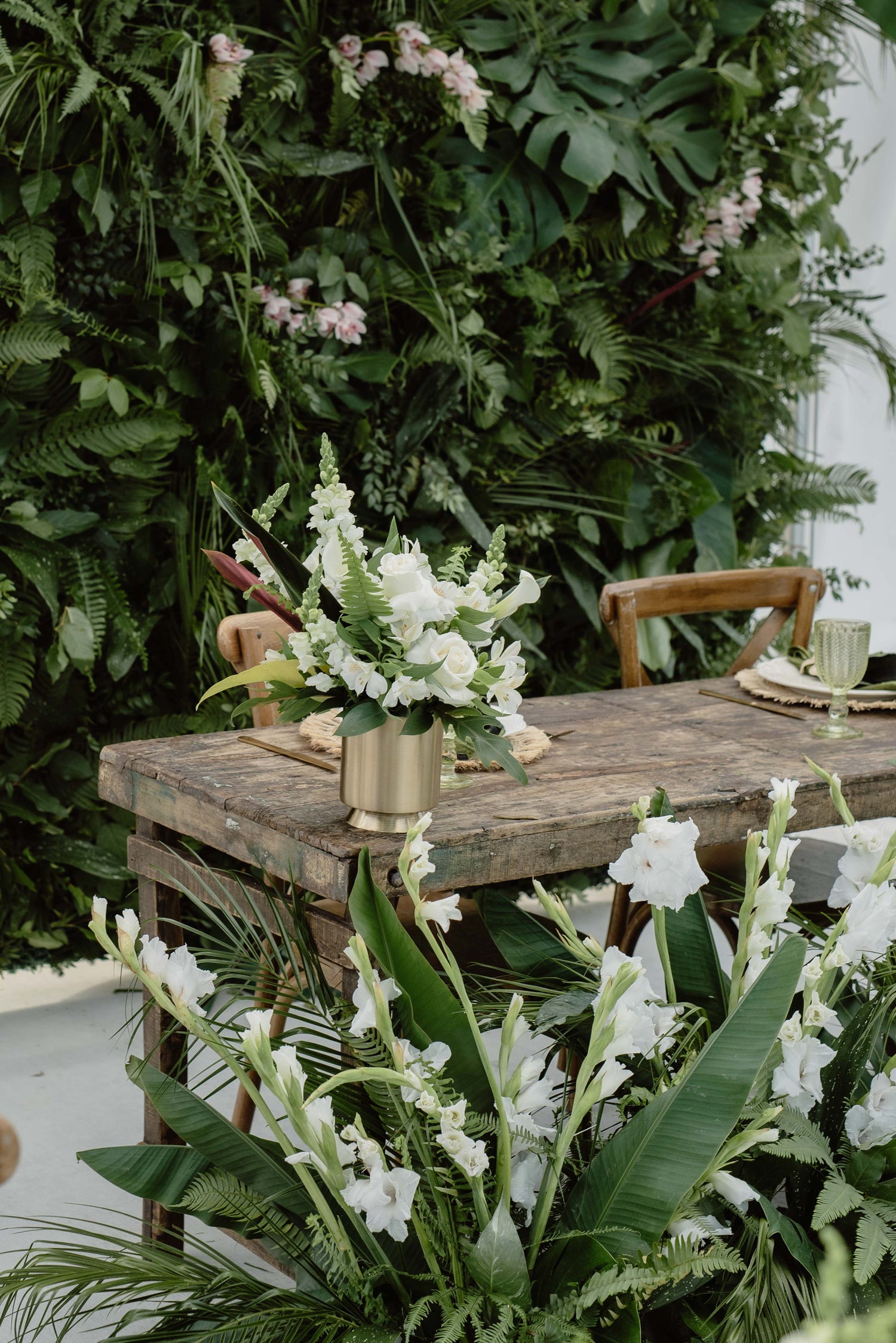 A rustic wooden table styled with white flowers in a gold vase, set against a lush green foliage wall backdrop.